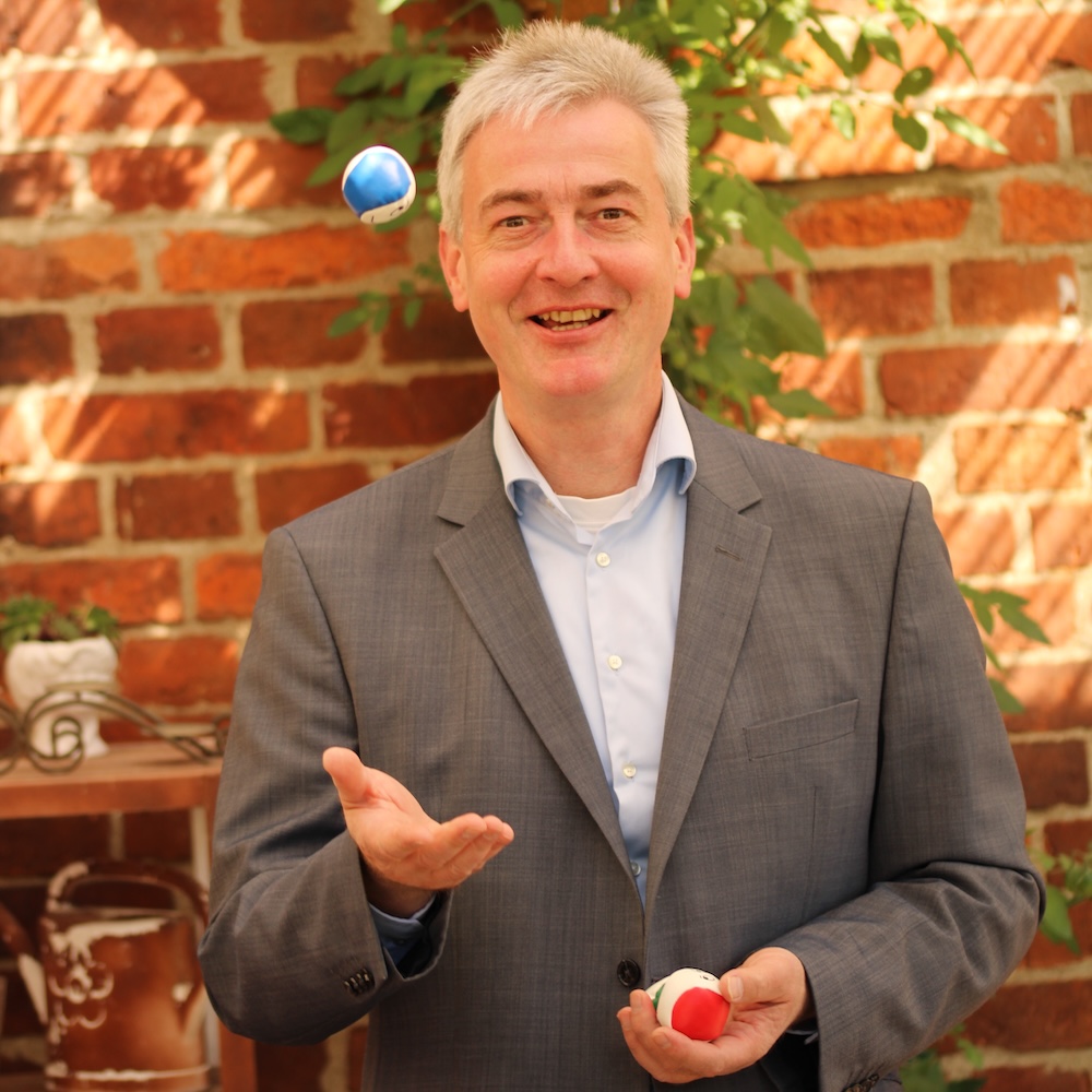 Das Foto zeigt ein Portraits von Riccardo Freiheit, der freundlich in die Kamera schaut und mit Bällen jongliert. Im Hintergrund eine rote Backsteinmauer und grüne Blätter.