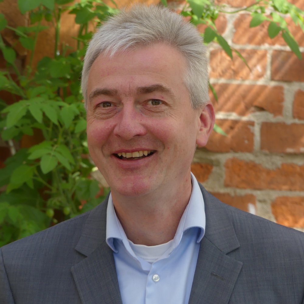 Das Foto zeigt ein Portraits von Riccardo Freiheit, der freundlich in die Kamera schaut. Im Hintergrund eine rote Backsteinmauer und grüne Blätter.